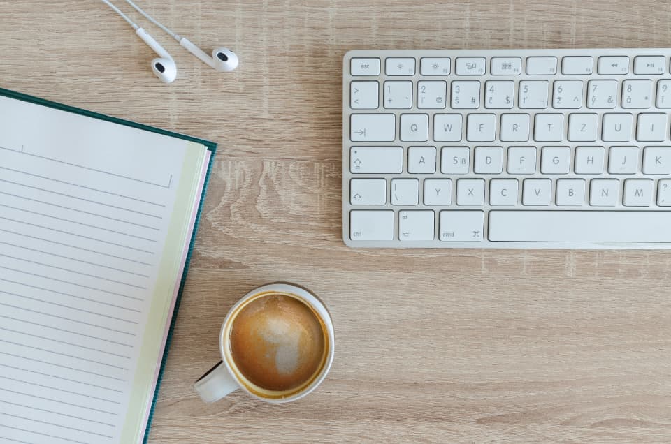Schreibtisch mit offenem Notizbuch, Tastatur und einer Tasse Kaffee