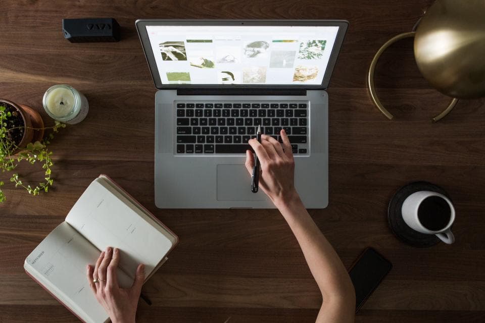 Blick von oben auf einen Holztisch: eine Hand am Laptop, die andere auf einem offenen Kalenderbuch, daneben eine Tasse Kaffee