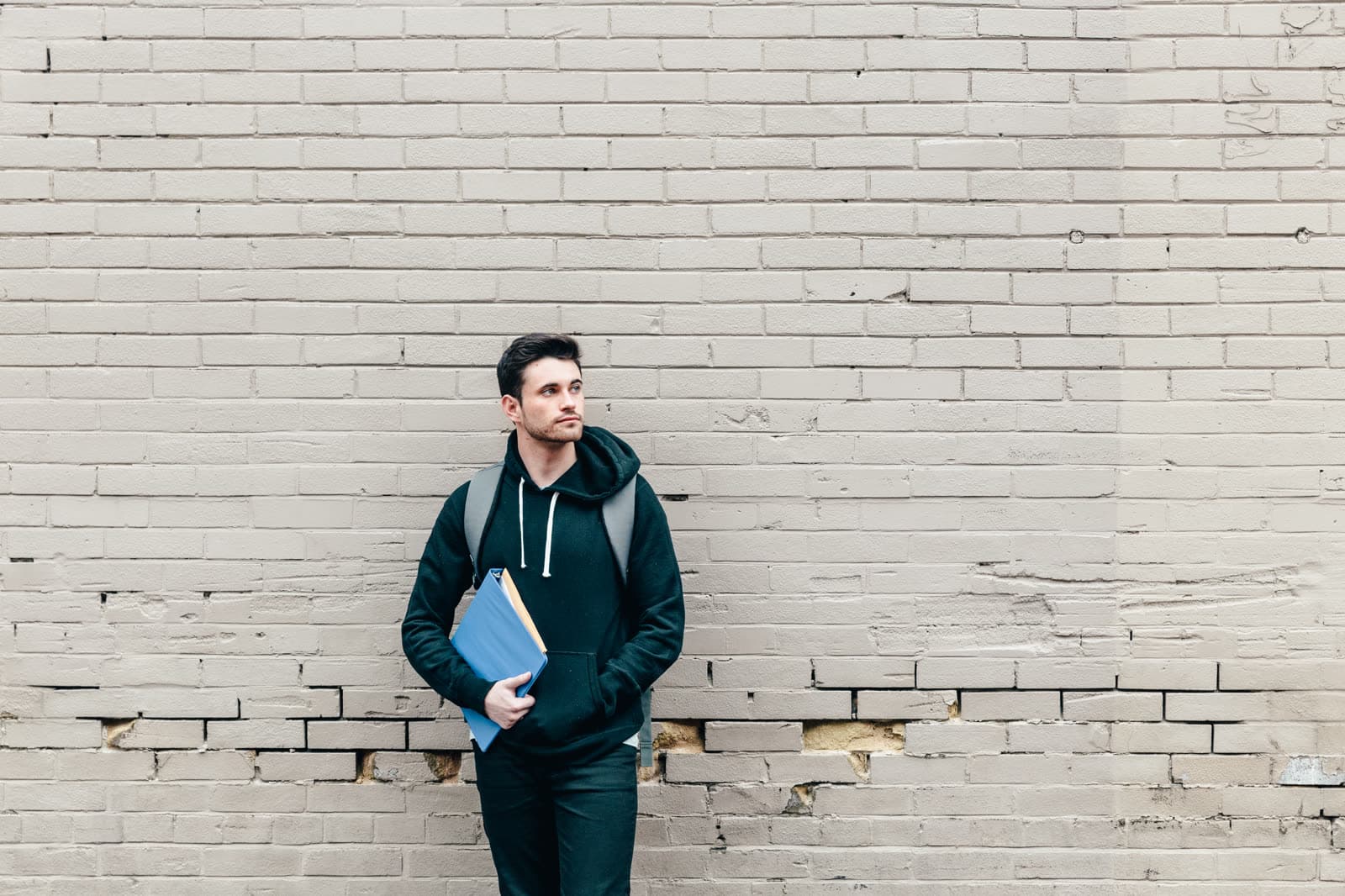 Junger Mann mit Rucksack und blauer Mappe lehnt an einer hellen Backsteinwand