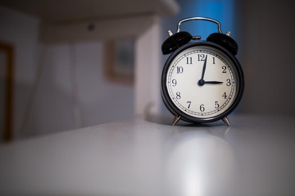 Schwarzer Wecker mit zwei Glocken auf einem hellen Tisch, die Zeiger stehen kurz nach drei Uhr
