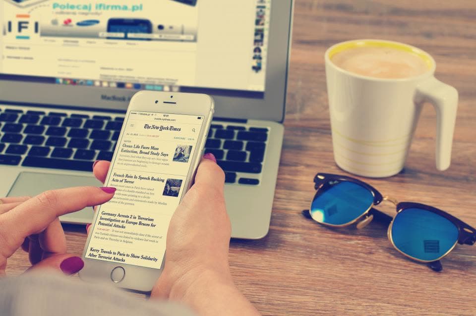 Smartphone mit Zeitungsartikel, Laptop, Kaffeetasse und Sonnenbrille auf einem Holztisch