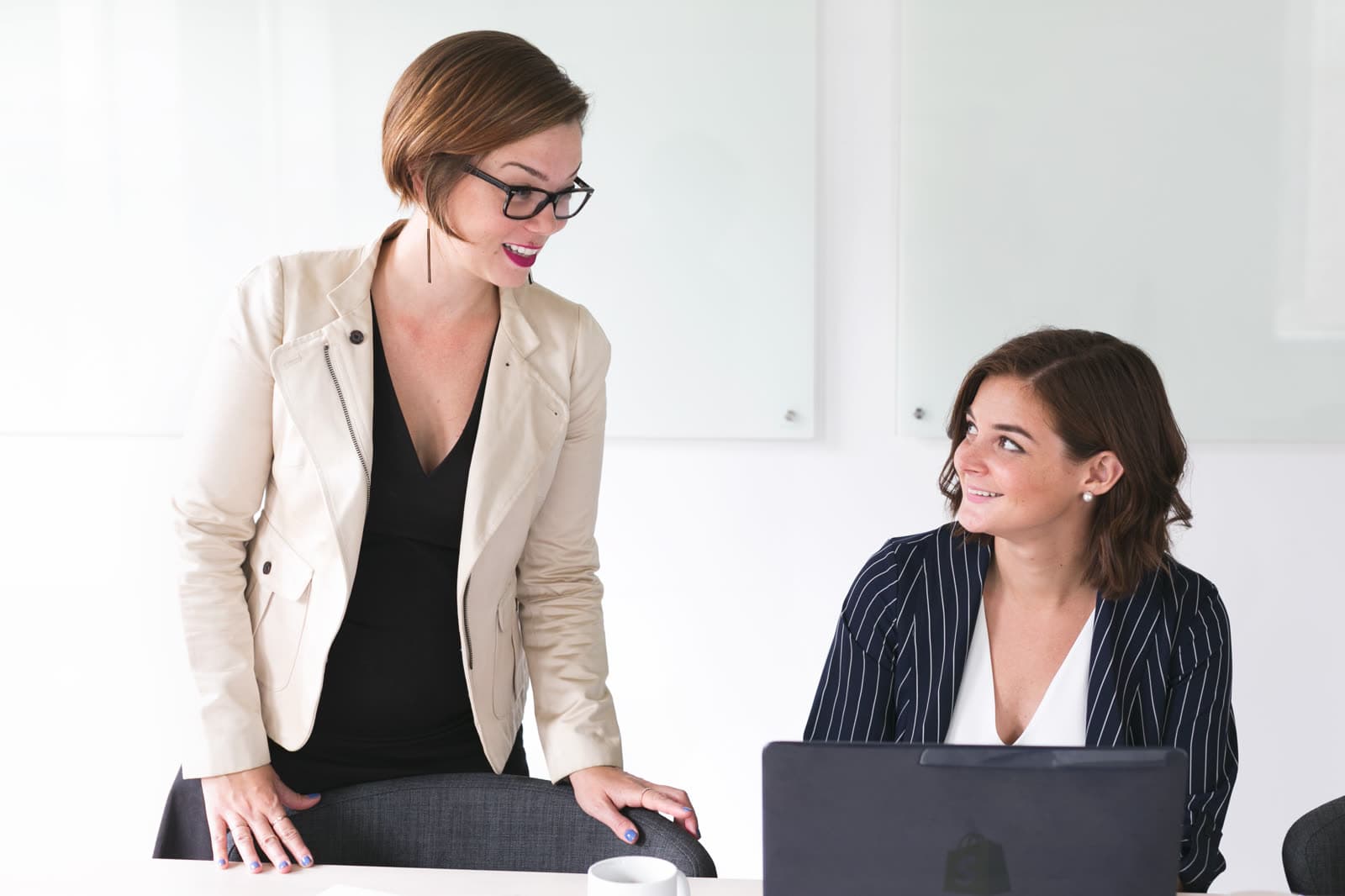 Zwei Frauen unterhalten sich in einem hellen Büro, eine steht, eine sitzt am Laptop